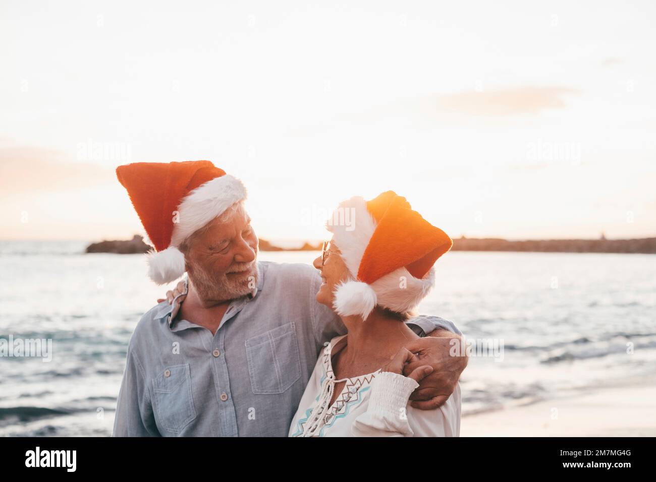 Old cute couple of mature persons enjoying and having fun together at the beach wearing christmas hats on holiday days. Walking on the beach with the sunset at the background at winter. Stock Photo