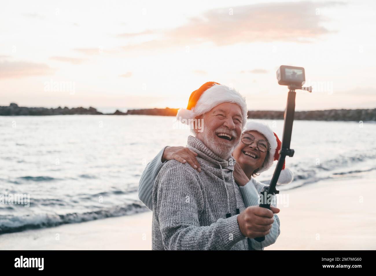 Portrait of two cute old persons having fun and enjoying together at the beach on christmas days at the beach wearing Christmas hats. Looking and holding a camera taking videos of vacations Stock Photo