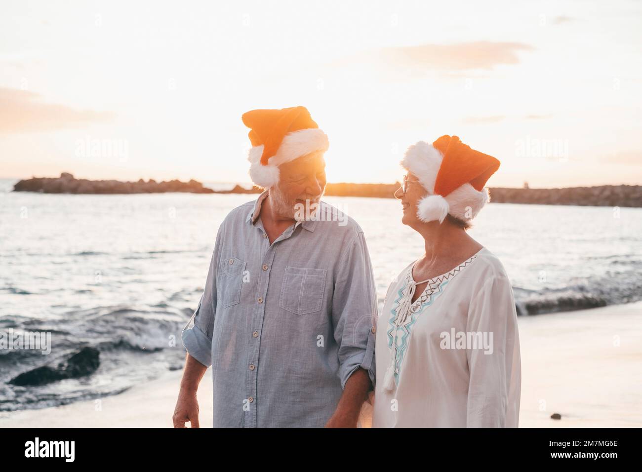 Old cute couple of mature persons enjoying and having fun together at the beach wearing christmas hats on holiday days. Walking on the beach with the sunset at the background at winter. Stock Photo