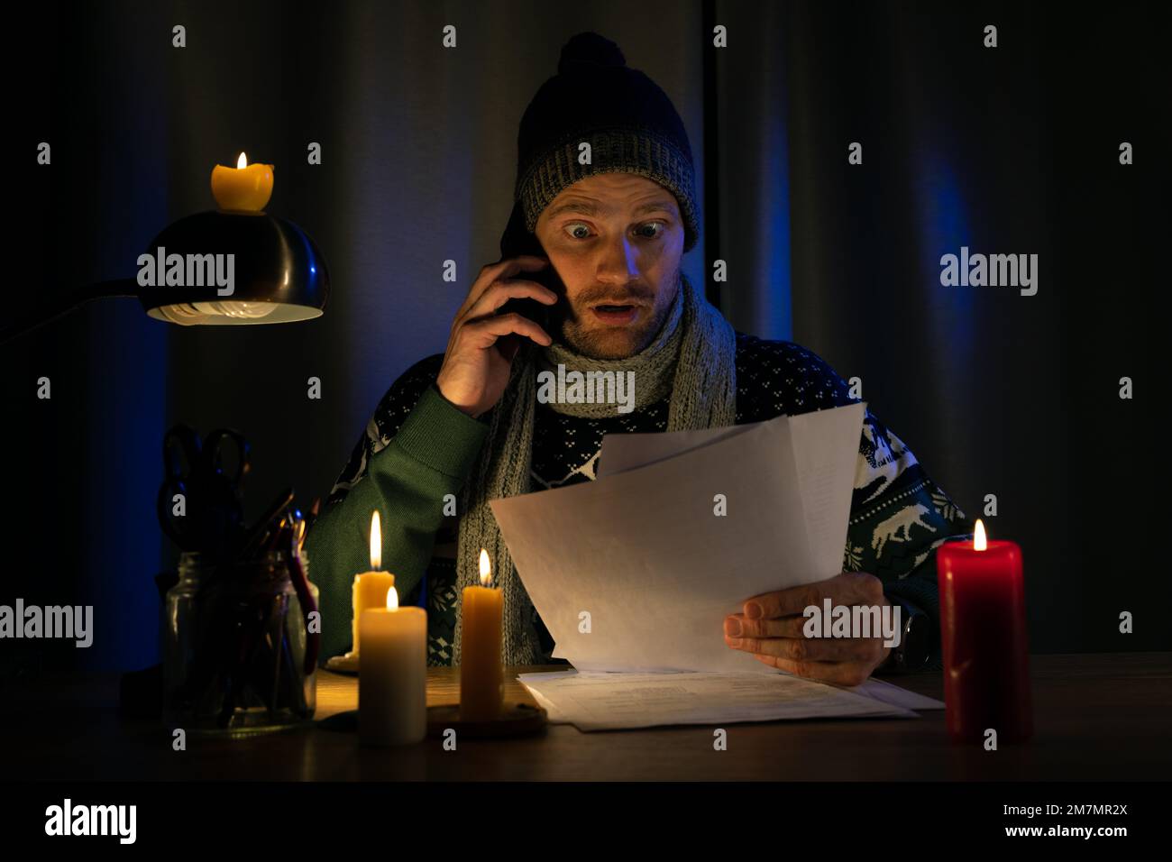 shocked man in candle light looking at high bills for electricity, gas heating in cold dark room at home. energy crisis, household expenses and inflat Stock Photo