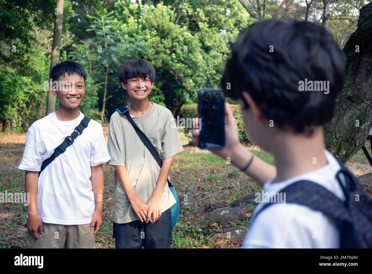A boy with a mobile phone taking a picture of two 13 year old boys side by side, outdoors in a park in summer. Stock Photo