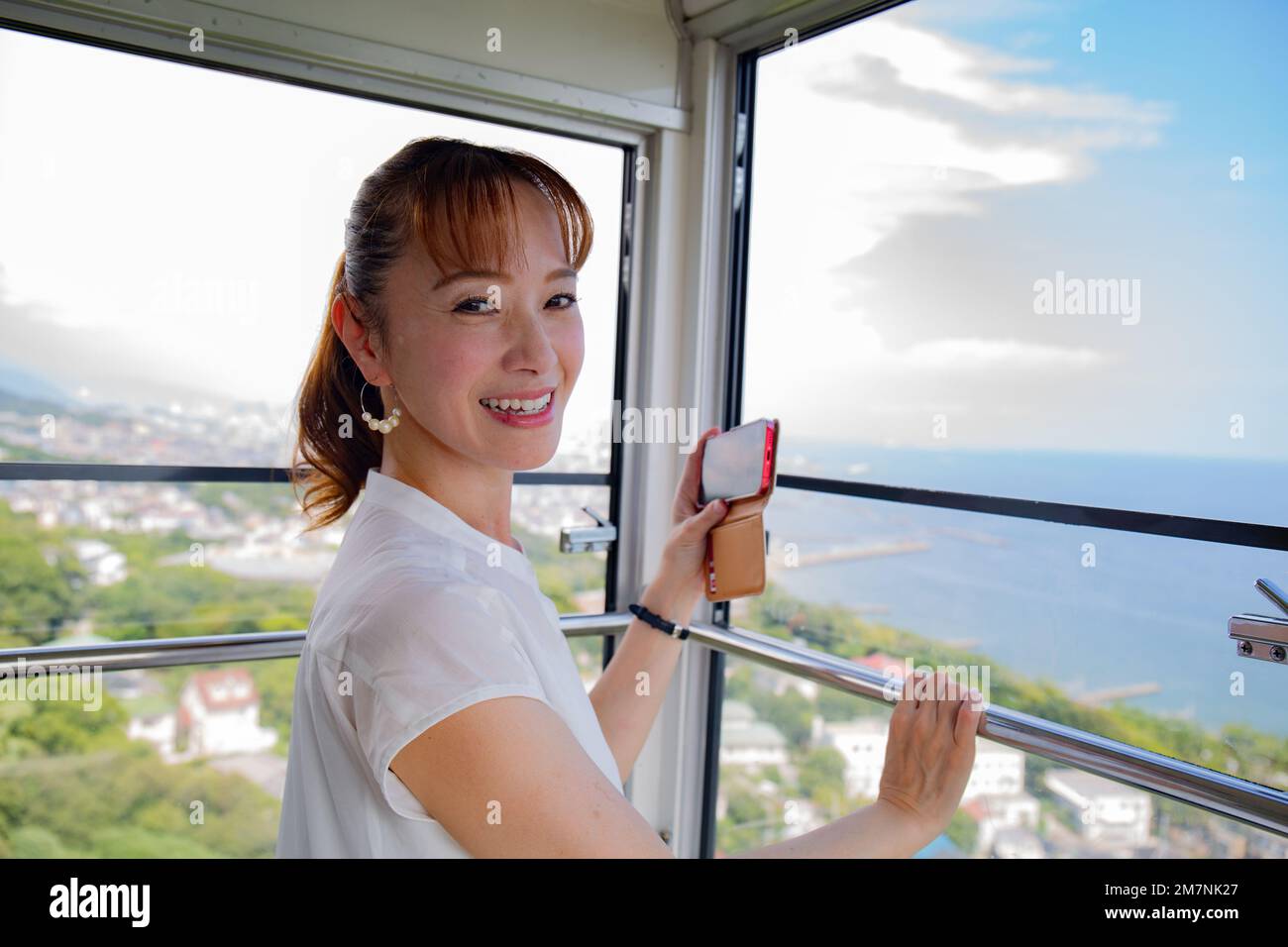 A mature Japanese woman using her mobile phone to take pictures from a cable car cabin of the city and landscape below. Stock Photo