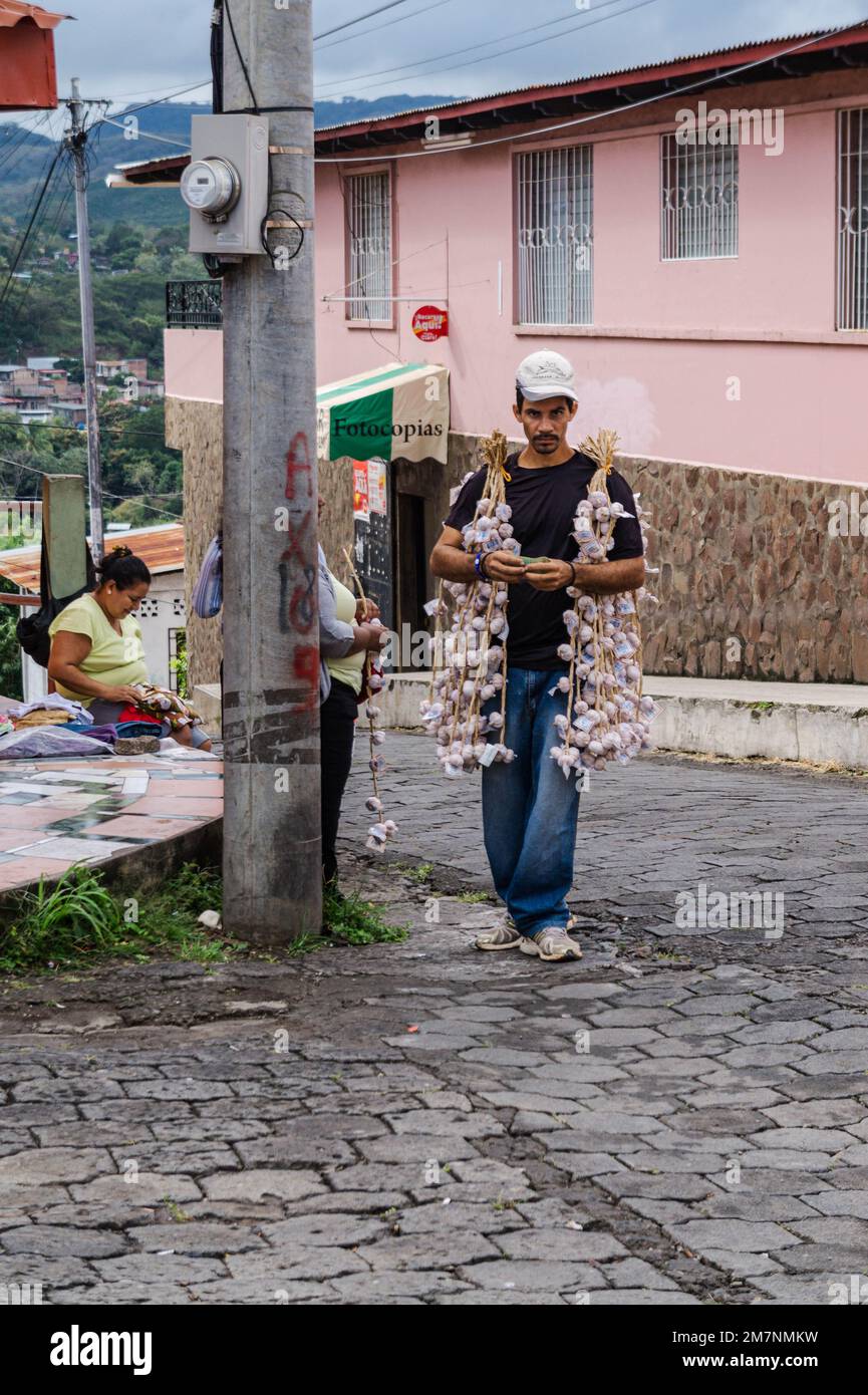 Garlic seller walks the streets of Boaco selling galic from braids of the bulbs draped around his shoulders,while another vendor sit on the sidewalk. Stock Photo