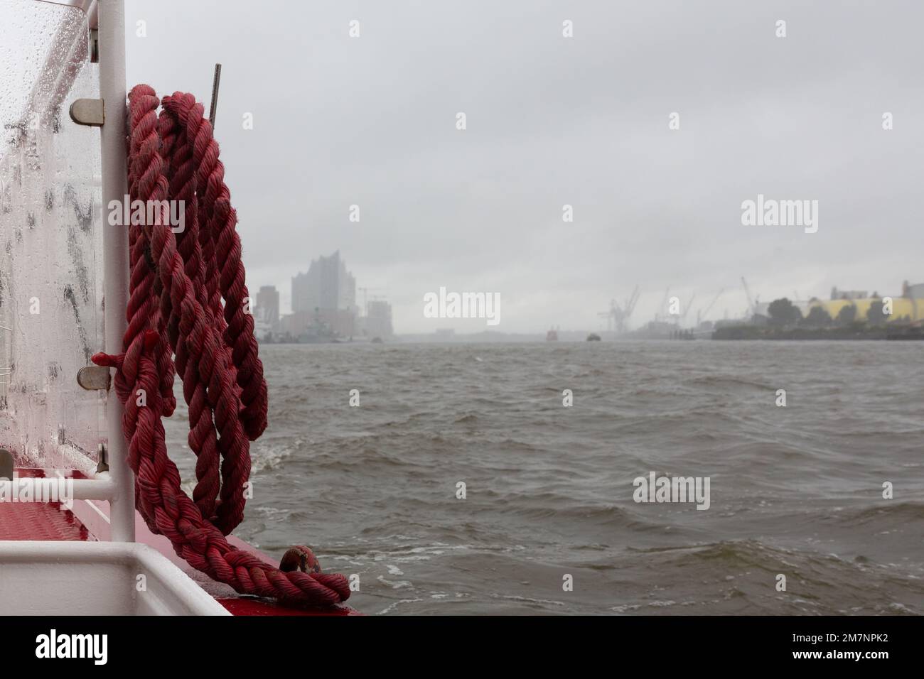A thick red rope hangs from the railing of a boat and gives a view of the entrance to the harbour in Hamburg. Stock Photo