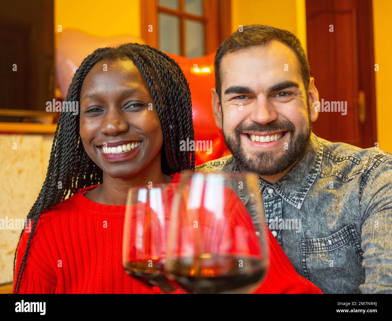 portrait of an interracial couple looking at camera laughing with glasses of red wine enjoying valentines day Stock Photo