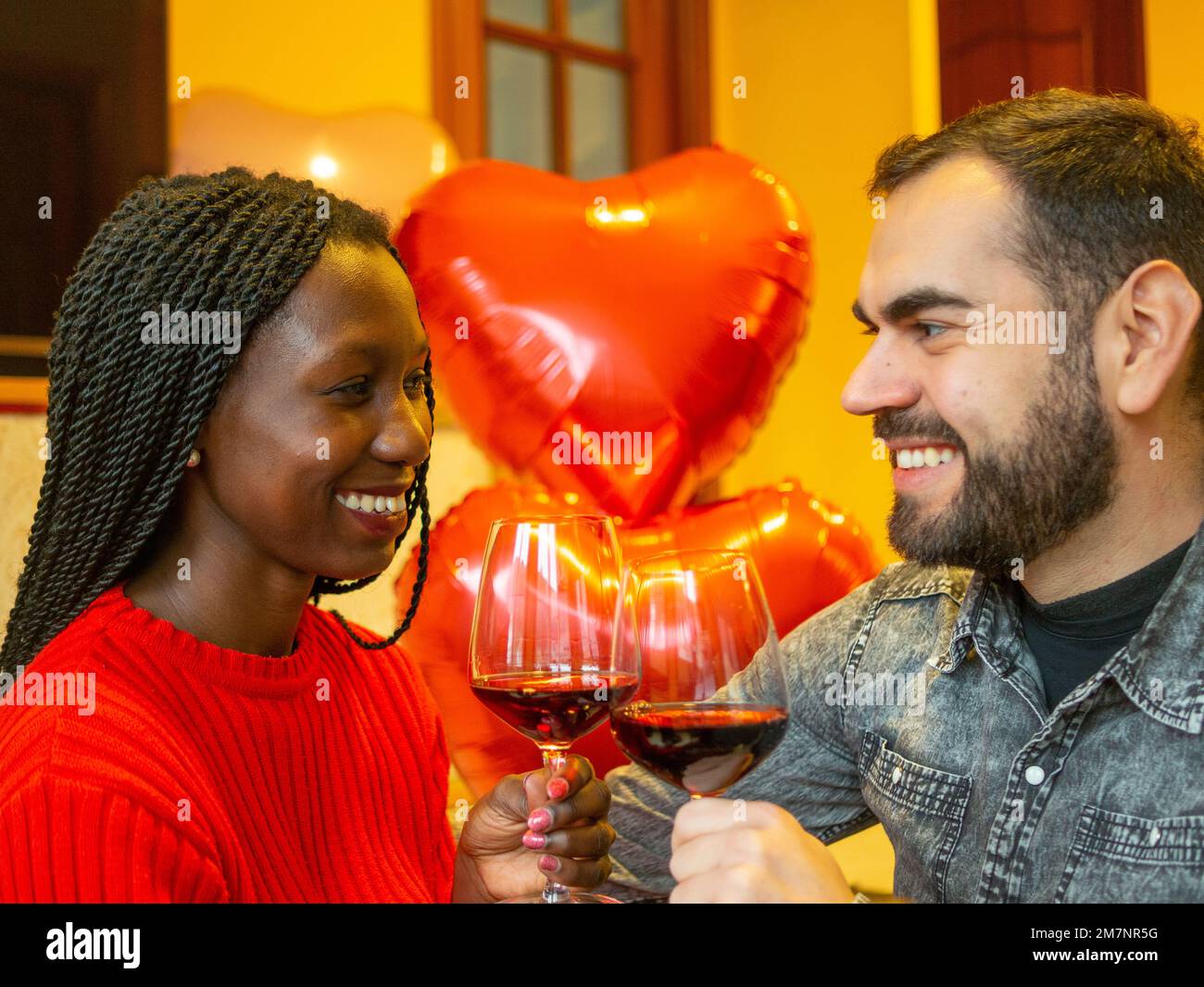 portrait of an interracial couple looking at each other laughing with glasses of red wine enjoying valentines day Stock Photo