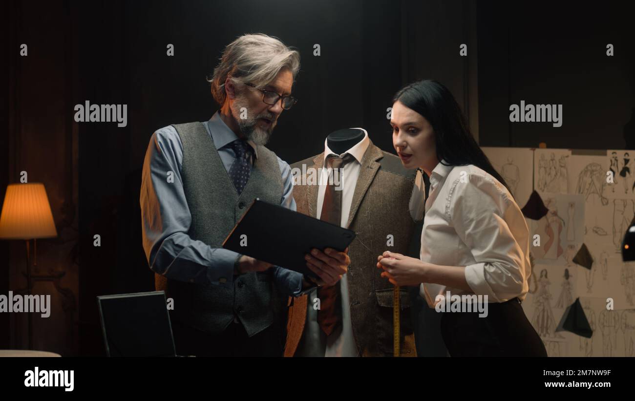 Male and female tailors work on business or wedding suit. Man draws sketch on tablet computer. Mannequin in tailored suit in designer atelier or tailoring studio. Fashion and hand craft concept. Stock Photo