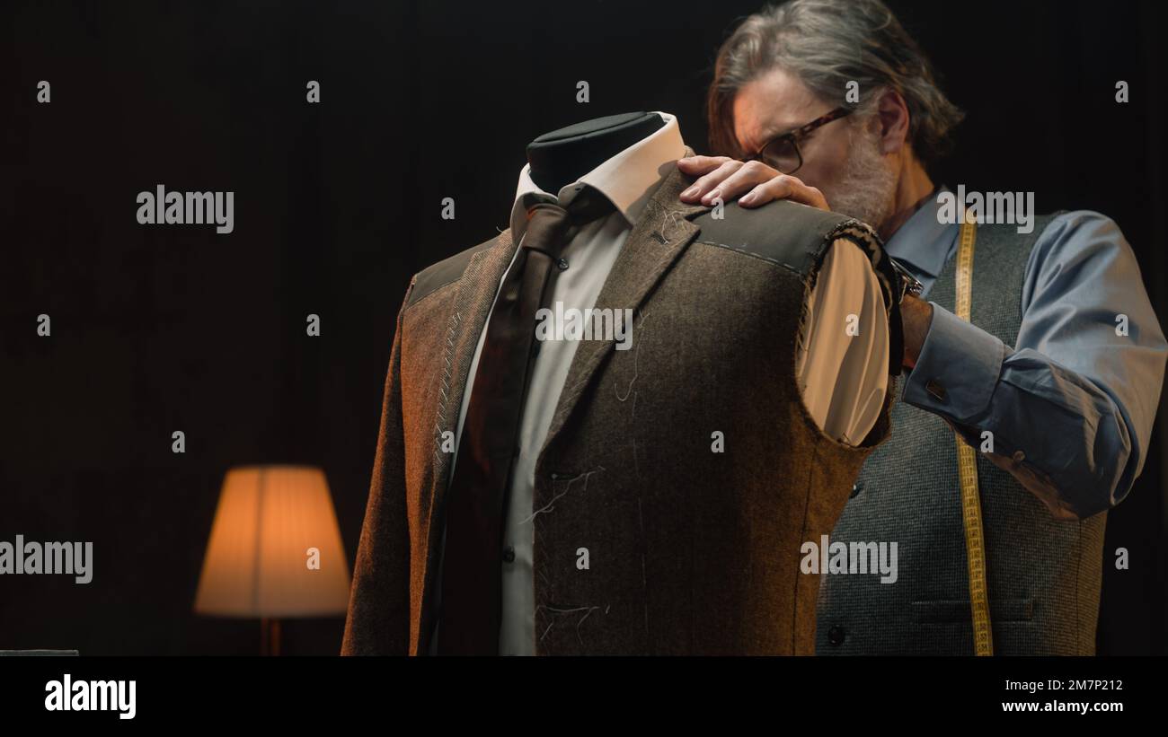 Male mature tailor works on elegant business or wedding suit. Mannequin with tailored shirt, tie and jacket in stylish luxury designer atelier or tailoring dim studio. Fashion and hand craft concept. Stock Photo