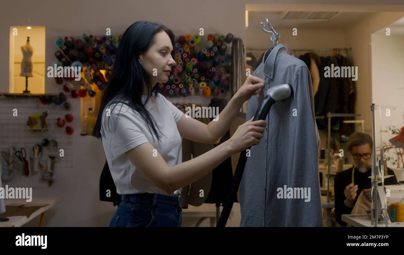 Seamstress irons new tailored shirt with electric hand steam generator in atelier workshop. Male tailor sews clothes on background. Concept of small business, tailoring industry and fashion. Stock Photo
