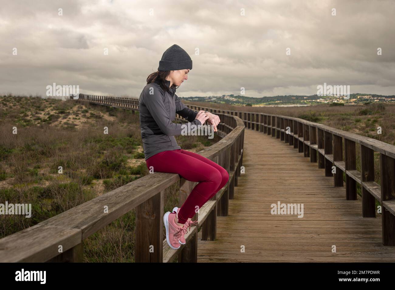 Female runner resting and checking her fitness tracker watch after running on a boardwalk. Stock Photo