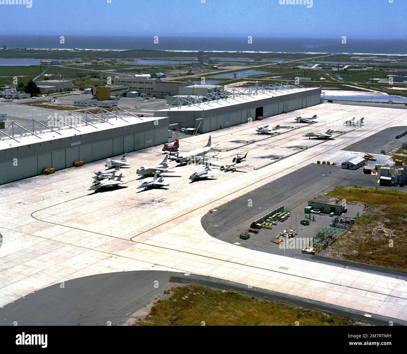 A view of the runway and hangars at Pacific Missile Test Center. Several aircraft are parked on the runway including an F/A-18 Hornet, F-14 Tomcat, P-3 Orion, F-4J Phantom II and a CH-46 Sea Knight helicopter. Base: Naval Air Station, Point Mugu State: California (CA) Country: United States Of America (USA) Stock Photo