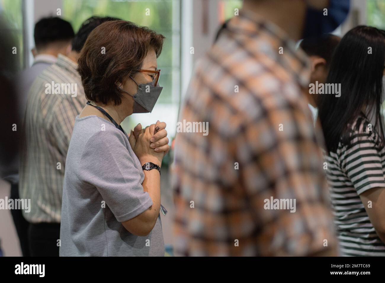 Religious Christian Asian woman with a face mask praying sincerely during a church worship service among a crowd. Stock Photo