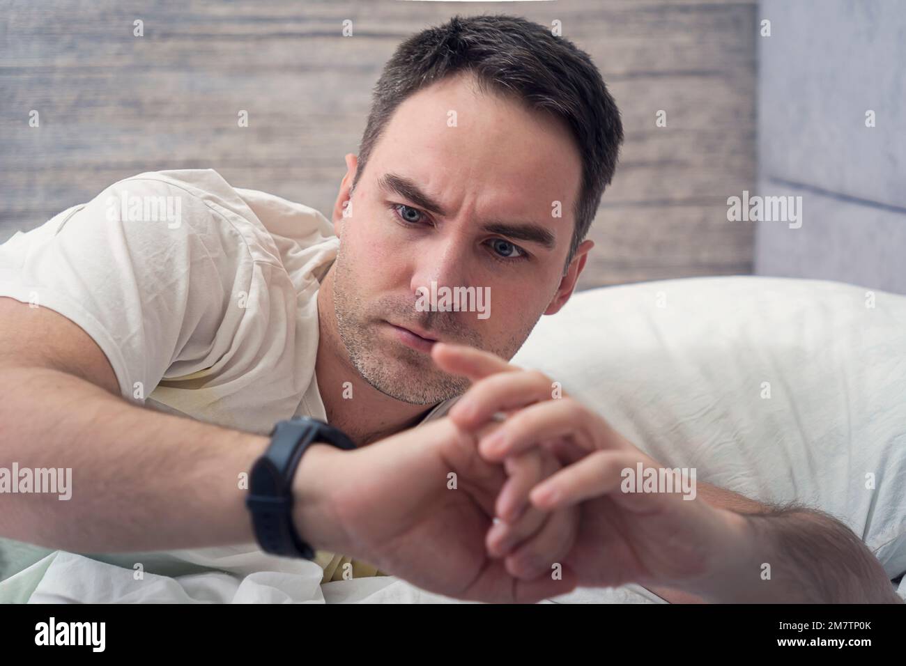 Stressed young man with emotional expression holding wrist watch and looking at time of alarm clock while lying on bed in bedroom. Surprise from the m Stock Photo
