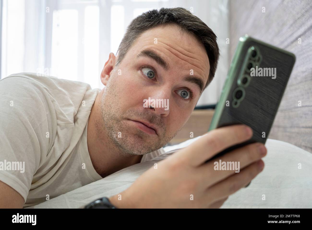 Man surprised to hear ringing of alarm smart phone, lies in white bed. Wake up shocked guy late for work in morning. Surprise from the message on smar Stock Photo