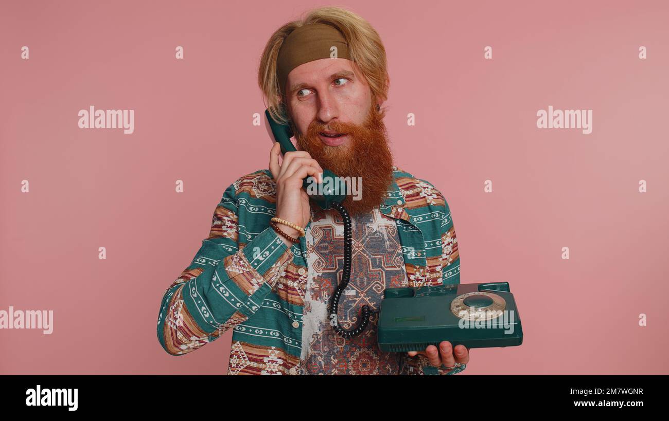Crazy hippie redhead bearded man in pattern shirt talking on wired vintage retro telephone of 80s, fooling making silly faces, unpleasant conversation. Hipster ginger guy boy on pink studio background Stock Photo