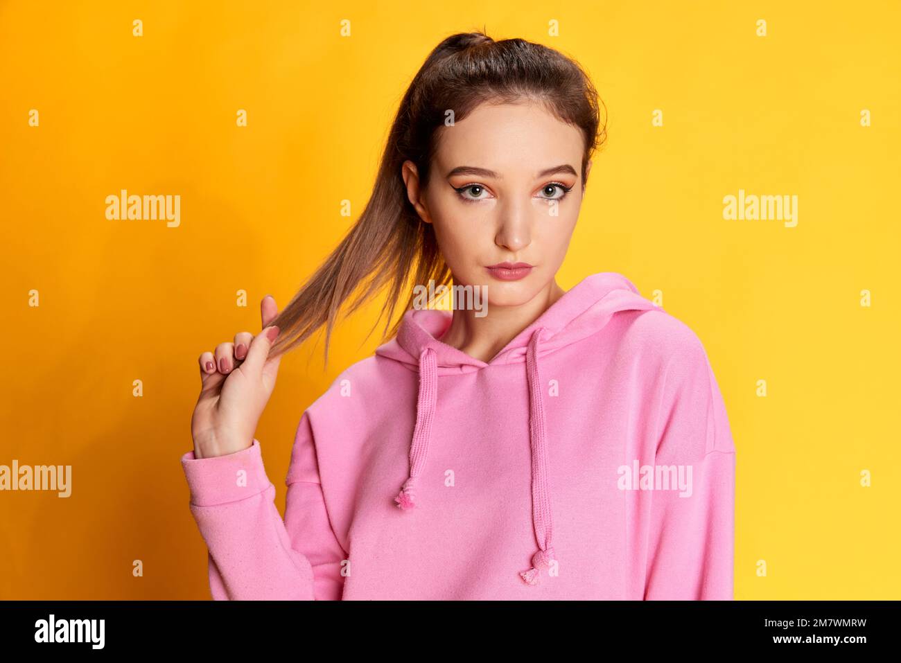 Portrait of young beautiful girl with ponytail posing in pink hoodie, looking at camera over yellow studio background. Concept of youth, emotions Stock Photo