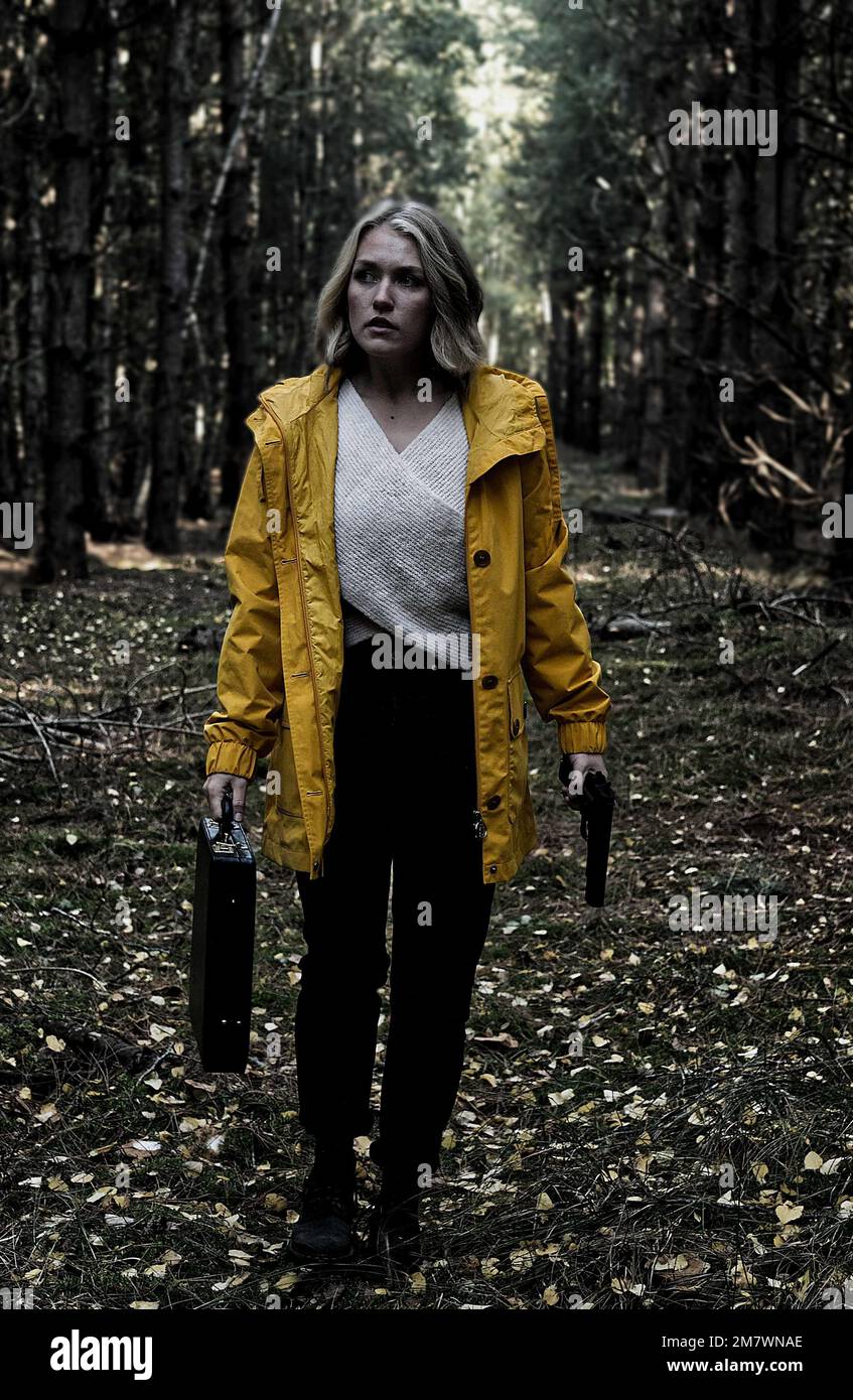 woman in yellow coat walking in a forest holding a gun (pistol) and a black briefcase. Spy, mystery thriller book cover style. Stock Photo