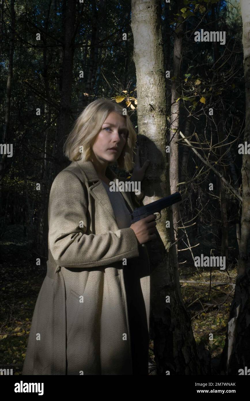 A young blonde woman wearing a big green coat, standing beside a tree and holding a pistol. thriller, adventure book cover style. Stock Photo