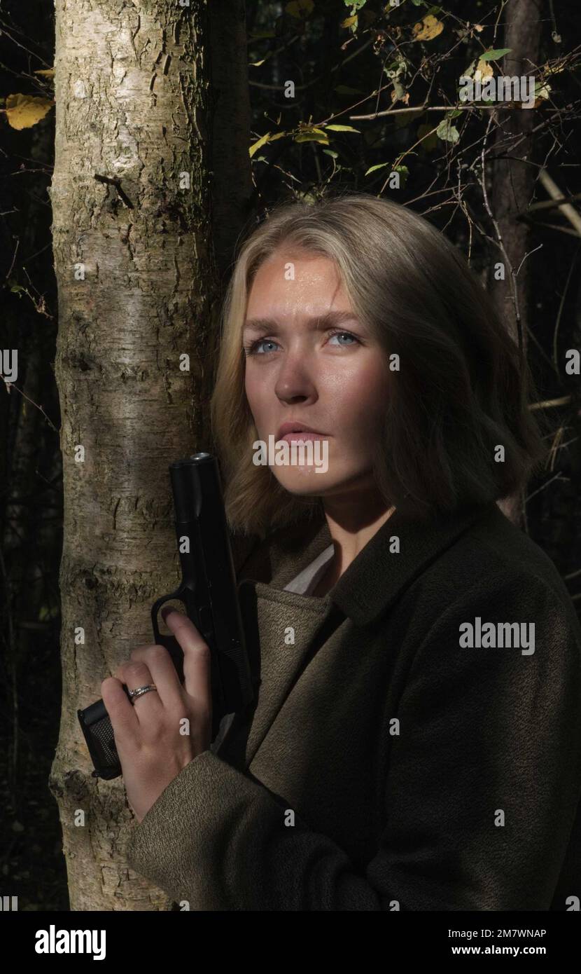 A young blonde woman wearing a big green coat, standing beside a tree and holding a gun (pistol)  thriller, adventure book cover style. Stock Photo