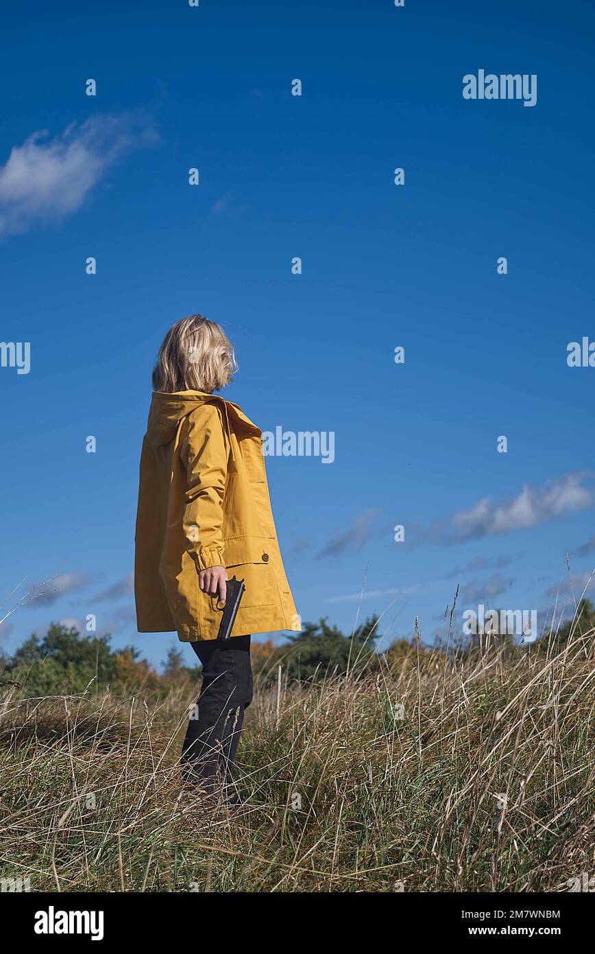 blonde woman with mid length hair and wearing a yellow jacket carrying a gun in the country side. thriller book cover style. Stock Photo