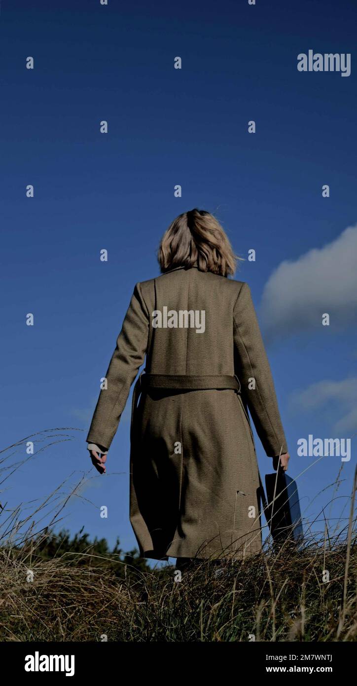 back view of young blonde woman walking in grass land,. Wearing a long green coat and carrying a brief case. Mystery, adventure book cover style. Stock Photo