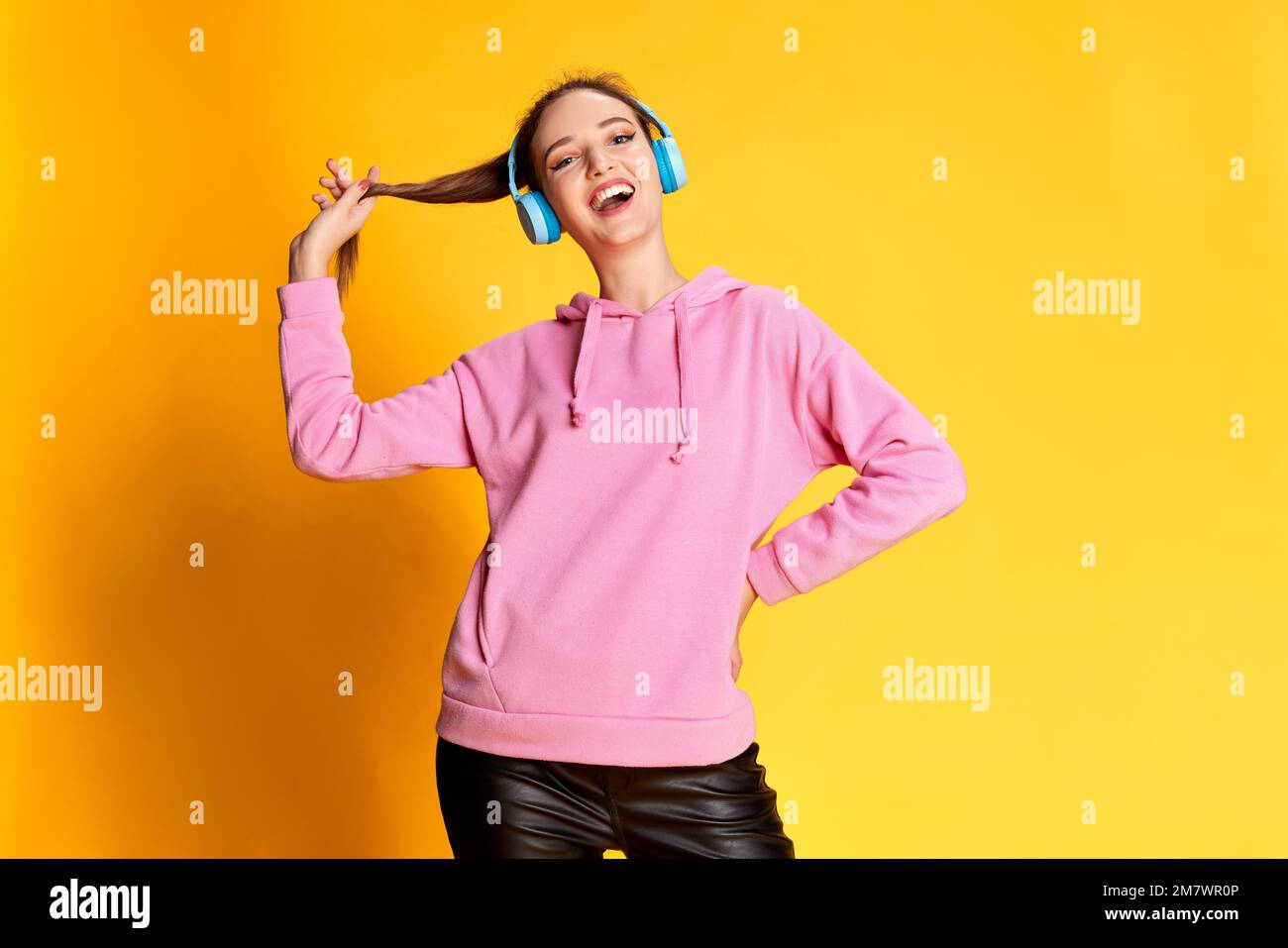 Portrait of young beautiful girl posing in pink hoodie, listening to music in headphones over yellow studio background. Concept of youth, emotions Stock Photo