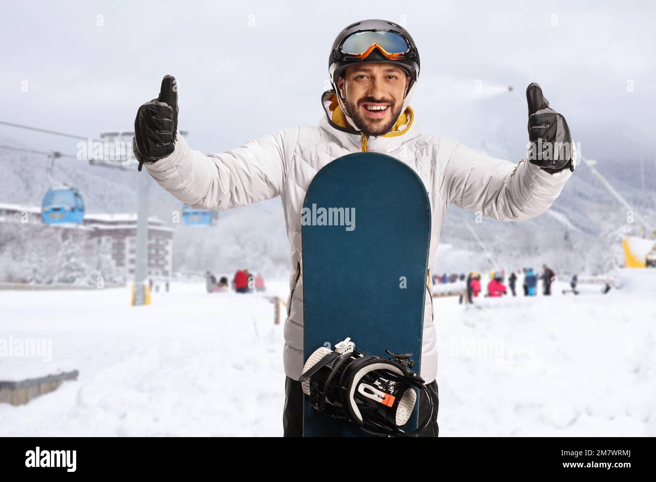 Young man with a snowboard gesturing with both thumbs up at a ski ...