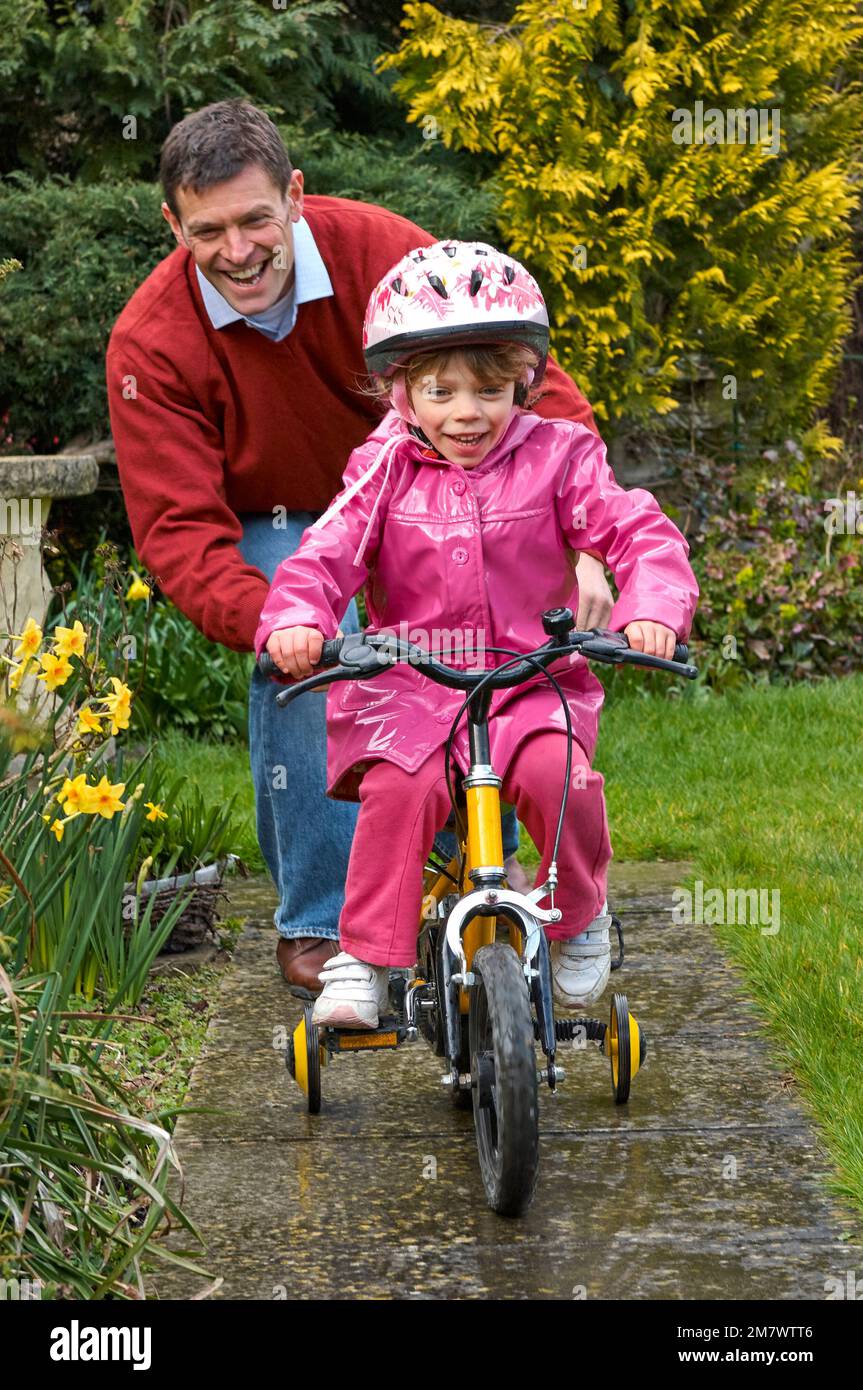 A man helping a young girl to ride her yellow bike with stabilisers on the pathway in a garden setting Stock Photo