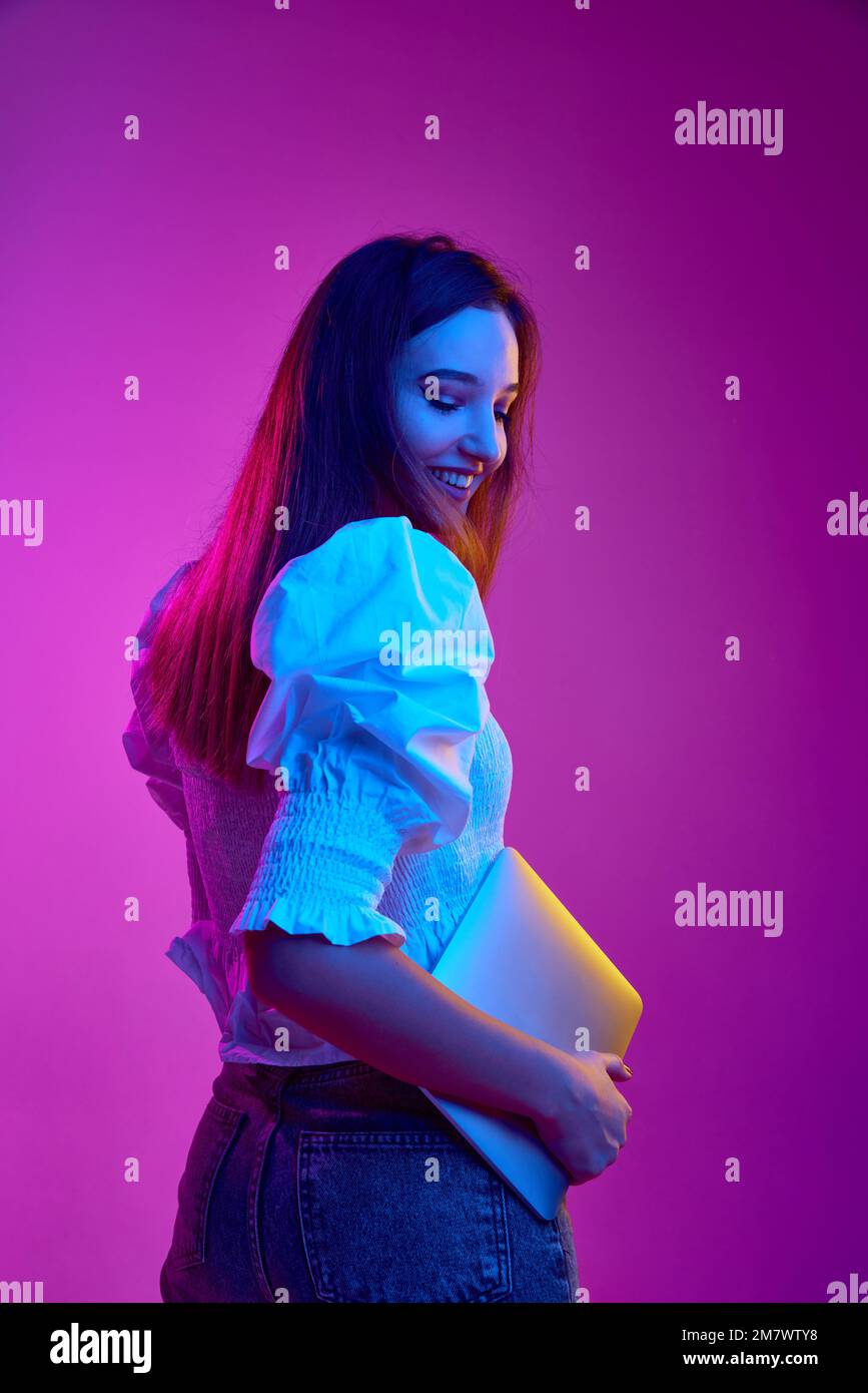 Portrait of young girl, student in white blouse posing with laptop over purple background in neon light. Online education. Concept of emotions, youth Stock Photo