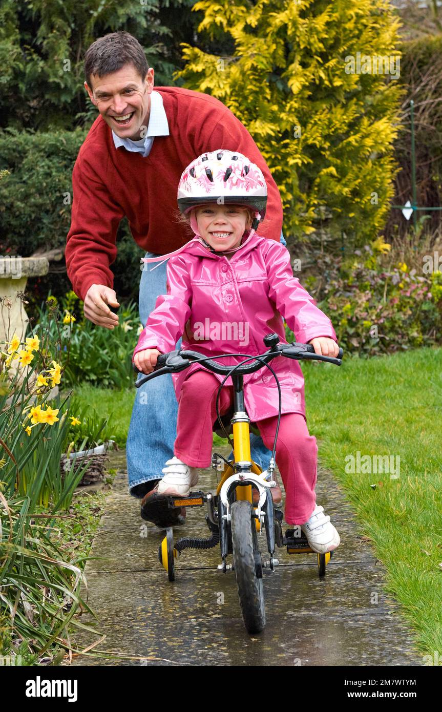 A man helping a young girl to ride her yellow bike with stabilisers on the pathway in a garden setting Stock Photo