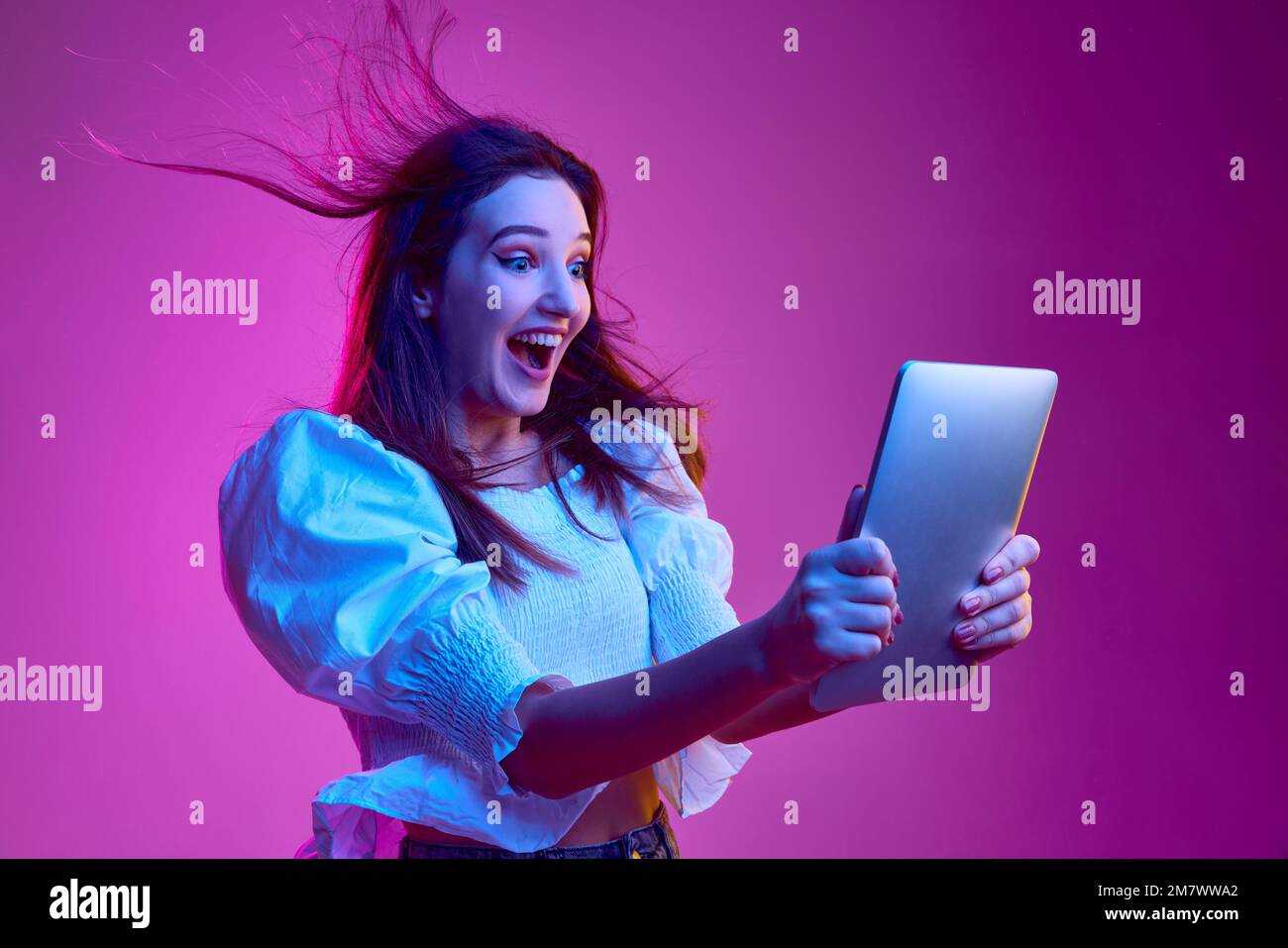 Portrait of young emotive girl cheerfully looking on tablet over pink background in neon light. Concept of emotions, youth, lifestyle Stock Photo