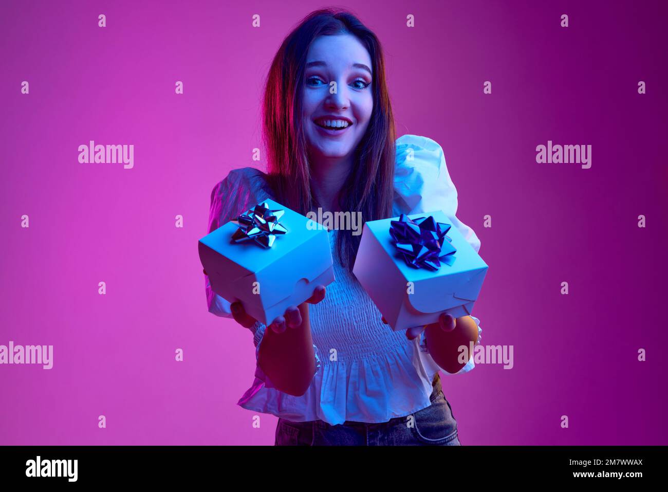 Portrait of young emotive girl holding two presents with happiness over pink background in neon light. Concept of emotions, youth, lifestyle Stock Photo