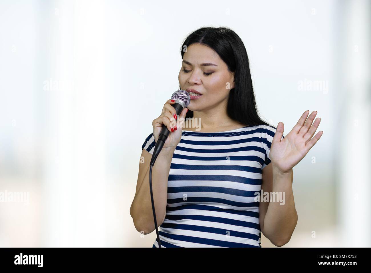 Young asianwoman is singing a song in a microphone standing indoors. Blurred bright background. Stock Photo
