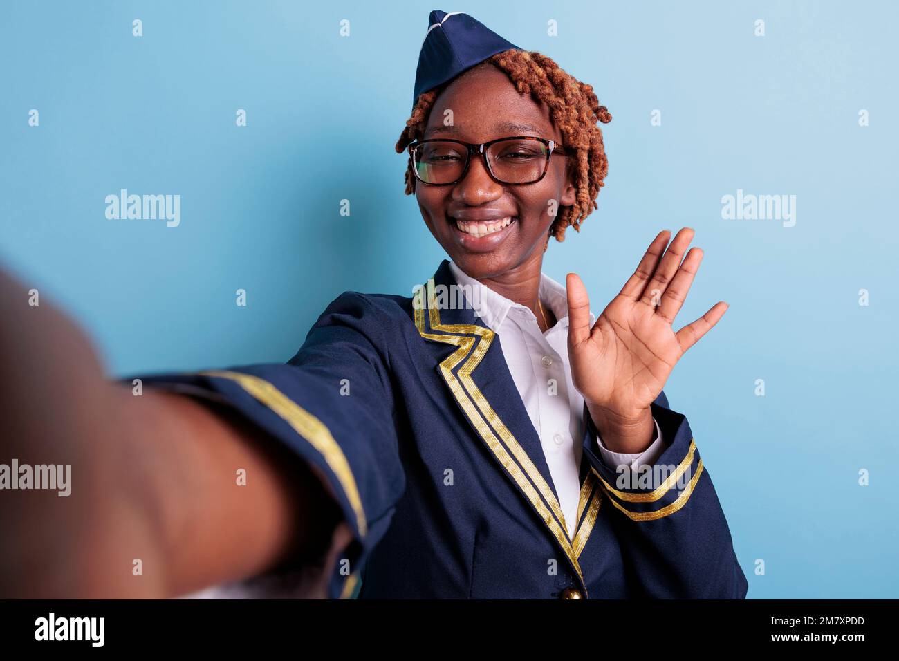 African american flight attendant waving happily in studio selfie. Aviation worker wearing glasses, afro woman taking self portrait with cellphone, smartphone. Stewardess portrait greeting. Stock Photo