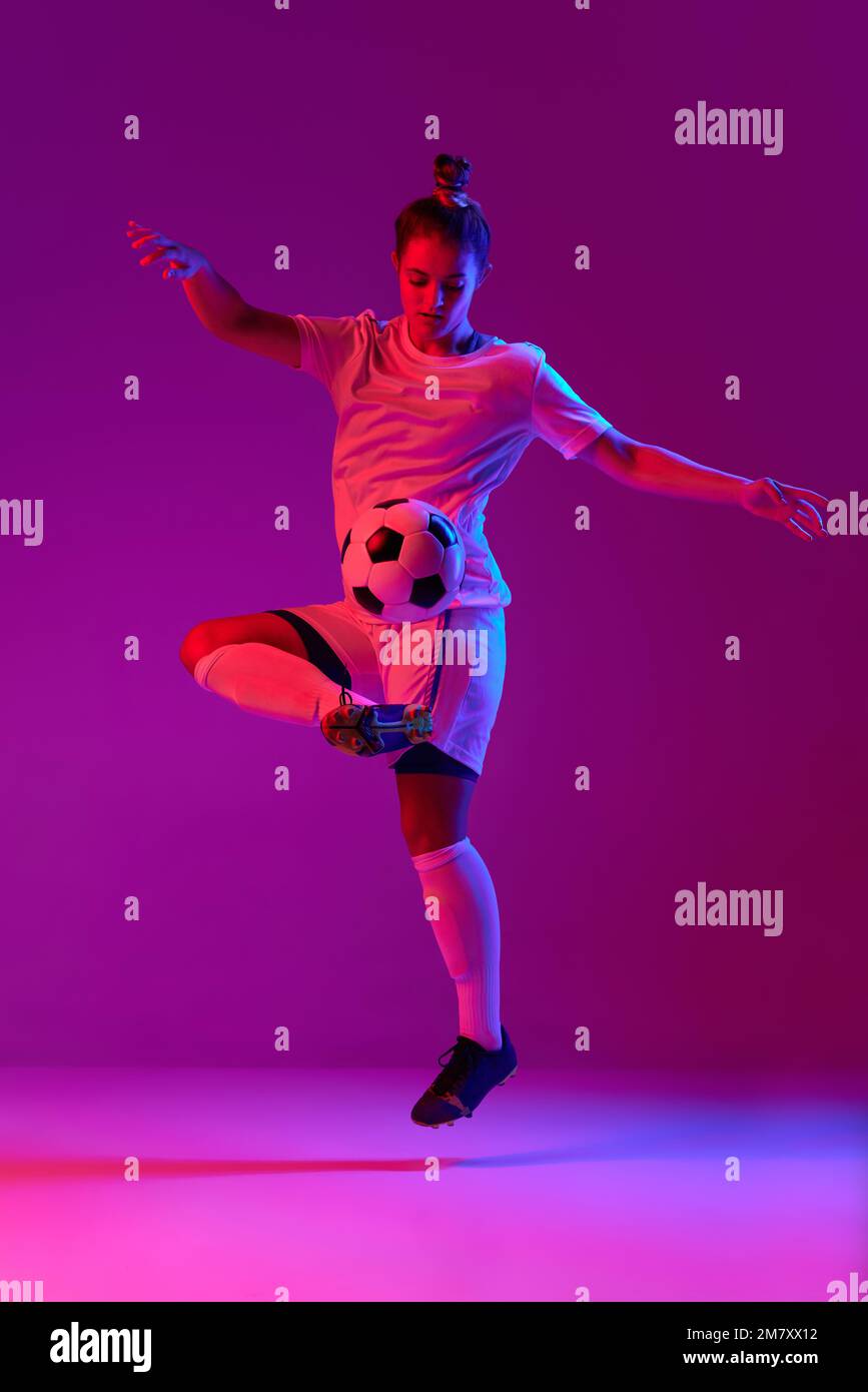 Young professional female football, soccer player in motion, training, playing over gradient pink background in neon light Stock Photo