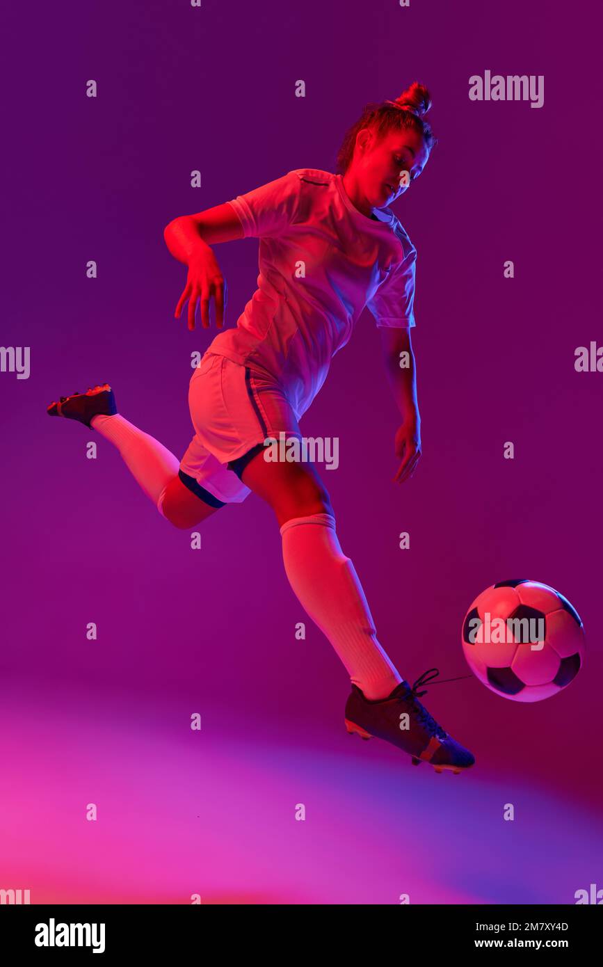 Young professional female football, soccer player in motion, training, playing over gradient pink background in neon light Stock Photo