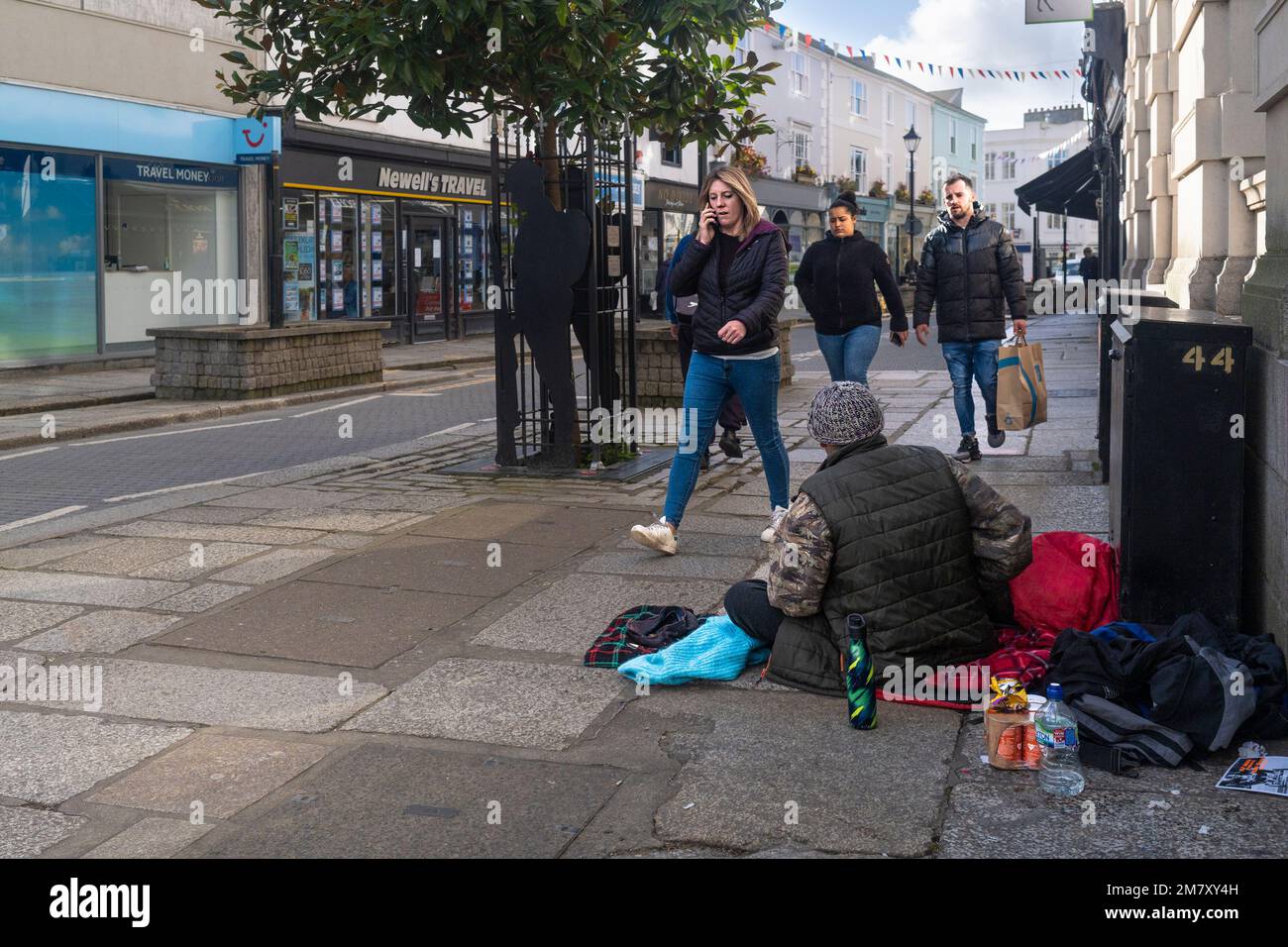 A homeless man male; sitting on the pavement sidewalk as people pedestrians walk past In Truro City centre in Cornwall in England in the UK. Stock Photo