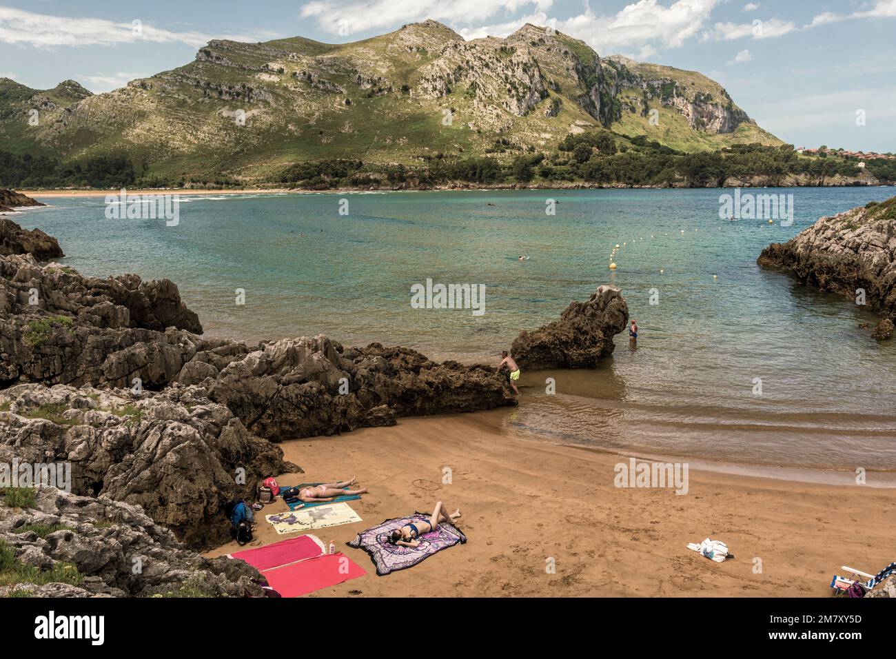 two women lying on the towel in bikinis on the sand of the small beach of the town of Islares, while other people bathe, Cantabria, Spain Stock Photo