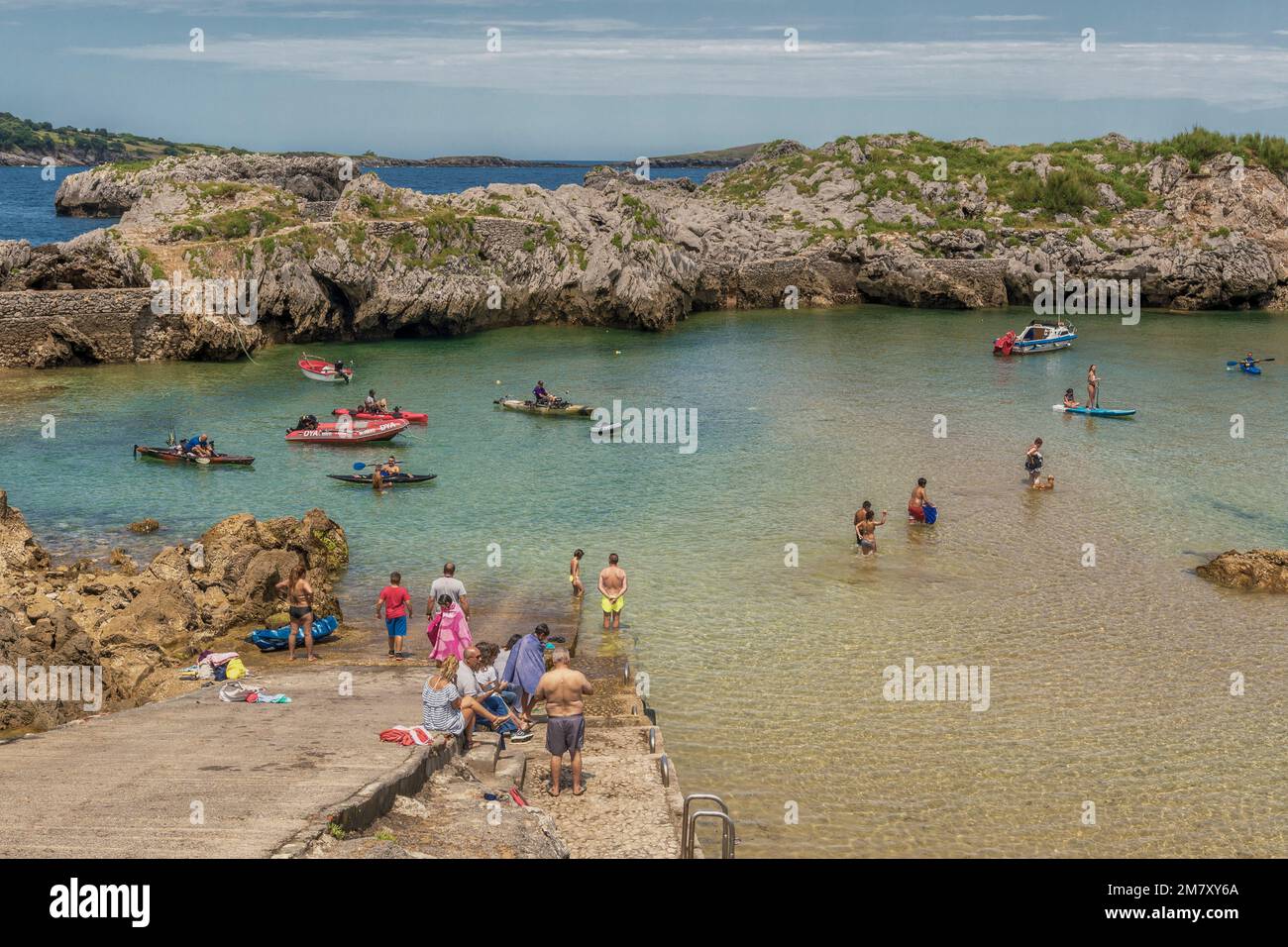 men, women, youth, boys and girls bathing, fishing in a canoe, paddle surfing and sitting sunbathing on a beach closed by rocks and stones in Spain. Stock Photo