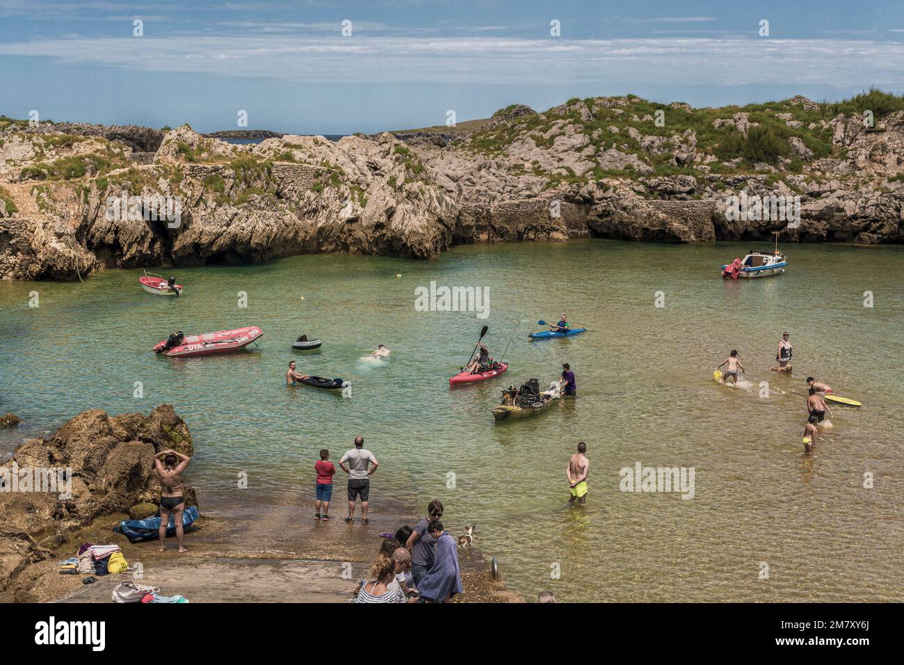 men, women, youth, boys and girls bathing, fishing in a canoe, paddle surfing and sitting sunbathing on a beach closed by rocks and stones in Spain. Stock Photo