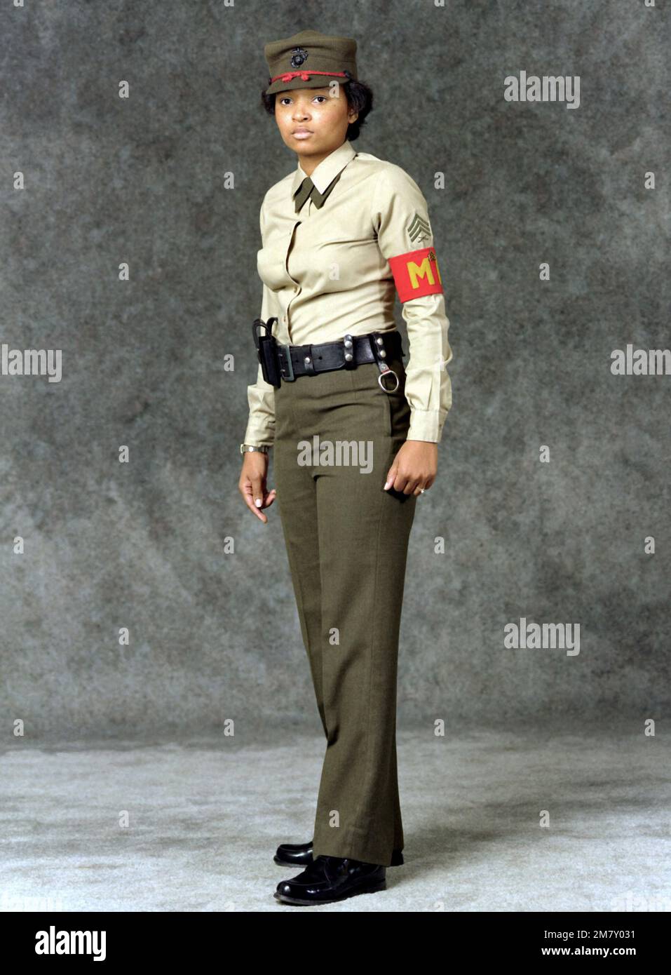 A woman Marine SGT. is dressed in a military police (MP) summer uniform.  She is wearing green slacks, a khaki shirt, a cap, an MP arm band and a  pistol belt. Base: