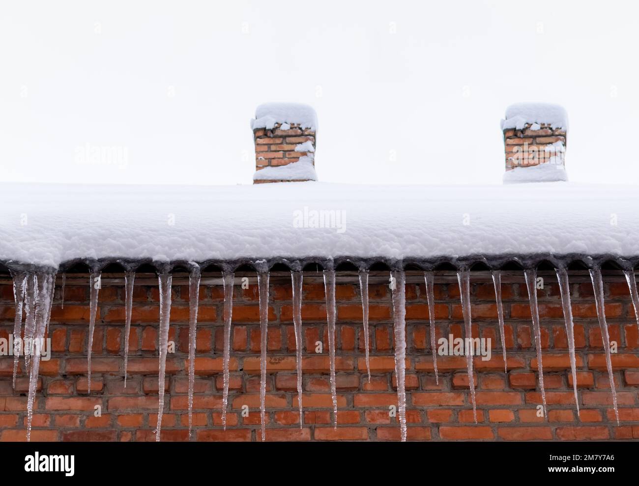 Snow-covered roof of a house with chimneys and icicles against red brick wall Stock Photo