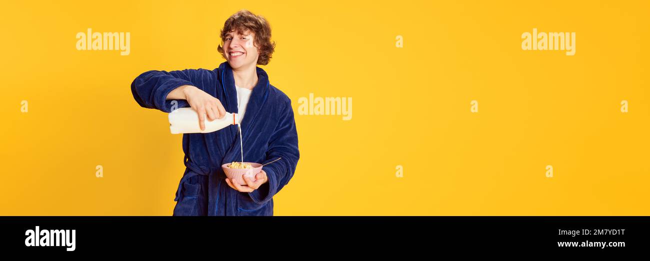 Smiling young man in bathrobe pouring milk into bowl isolated over