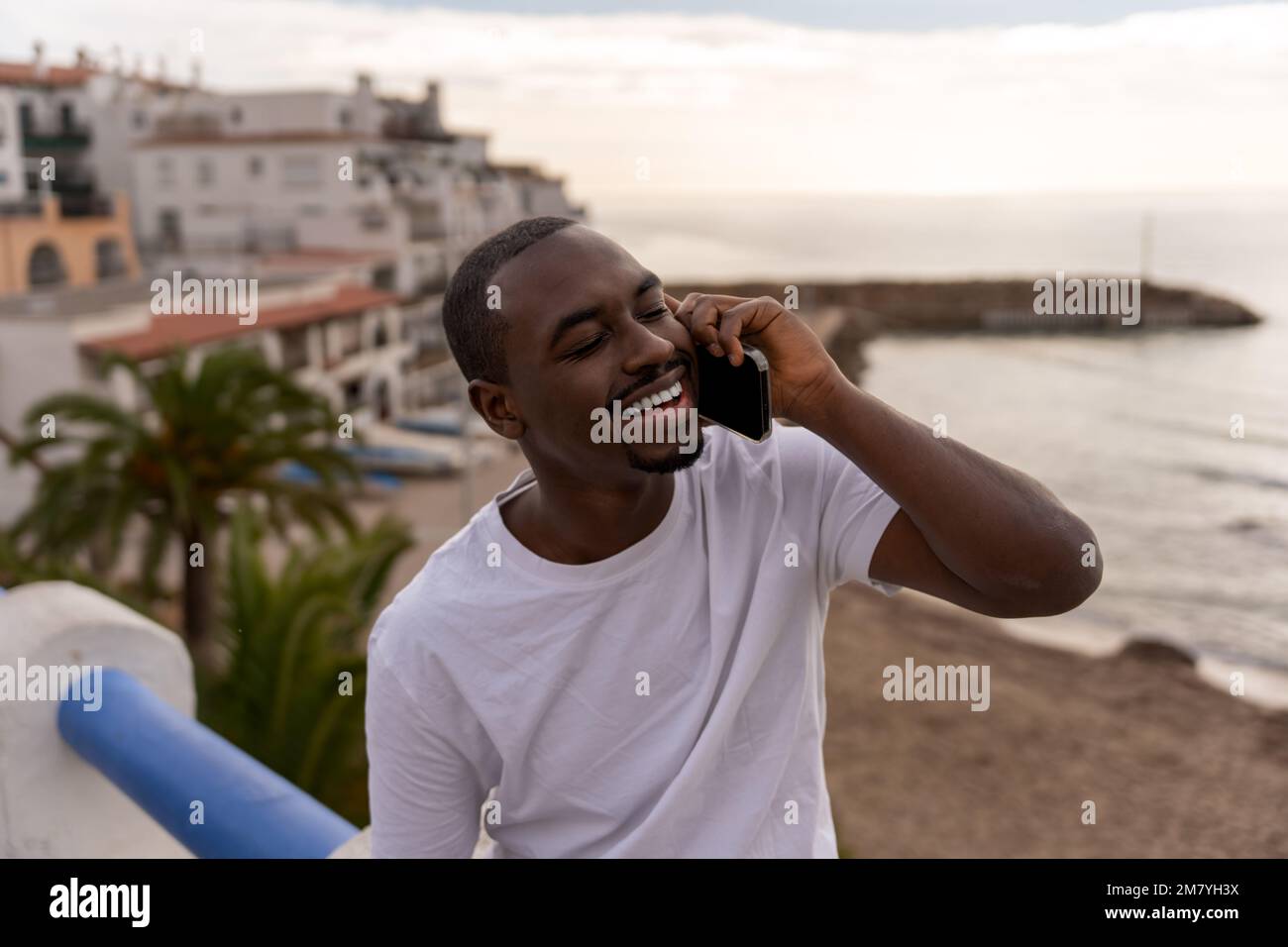 Positive African American male in casual clothes smiling and talking on smartphone while relaxing near sea during vacation Stock Photo