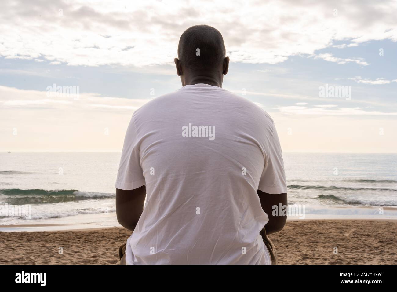 Low angle back view of unrecognizable African American male in casual white t shirt sitting on sandy beach and admiring waving sea Stock Photo