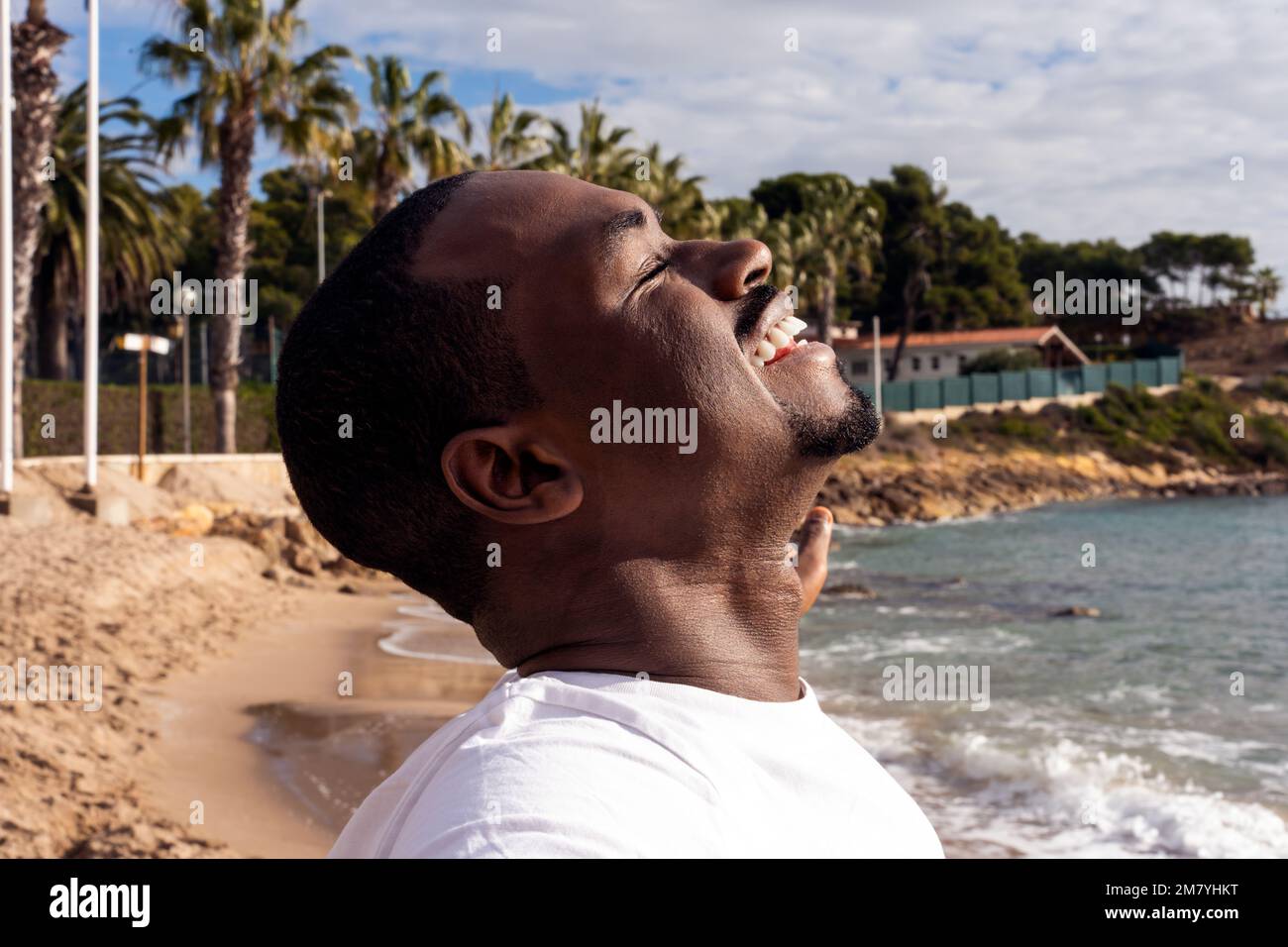 Happy black man in white t shirt spreading arms and smiling with closed eyes while breathing fresh air on sandy beach near waving sea Stock Photo