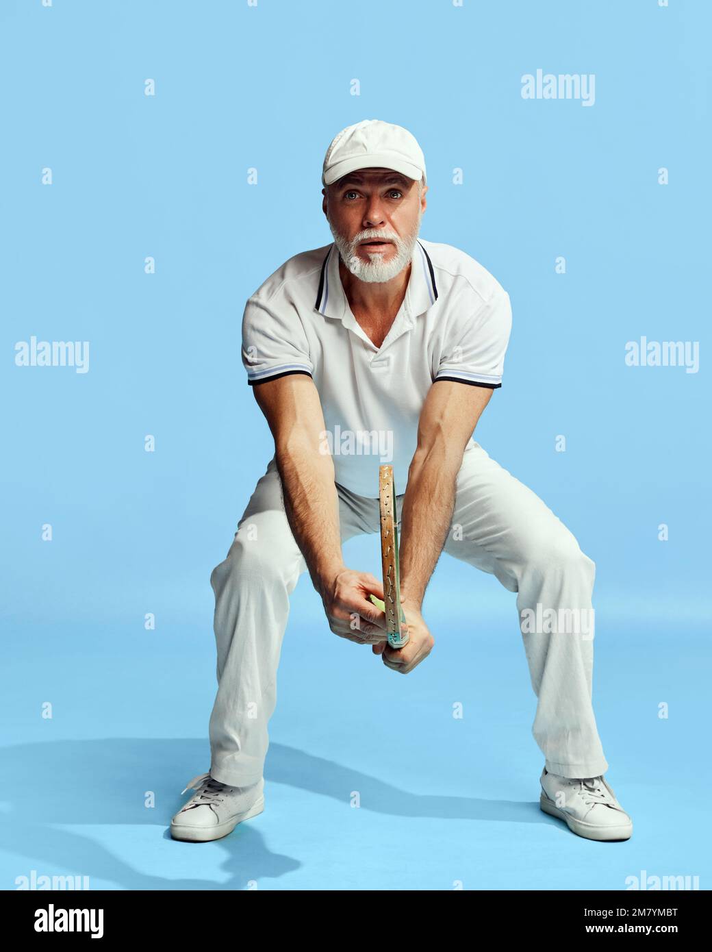 Concentration. Portrait of handsome senior man in stylish white outfit holding tennis racket over blue background. Concept of leisure activity, hobby Stock Photo