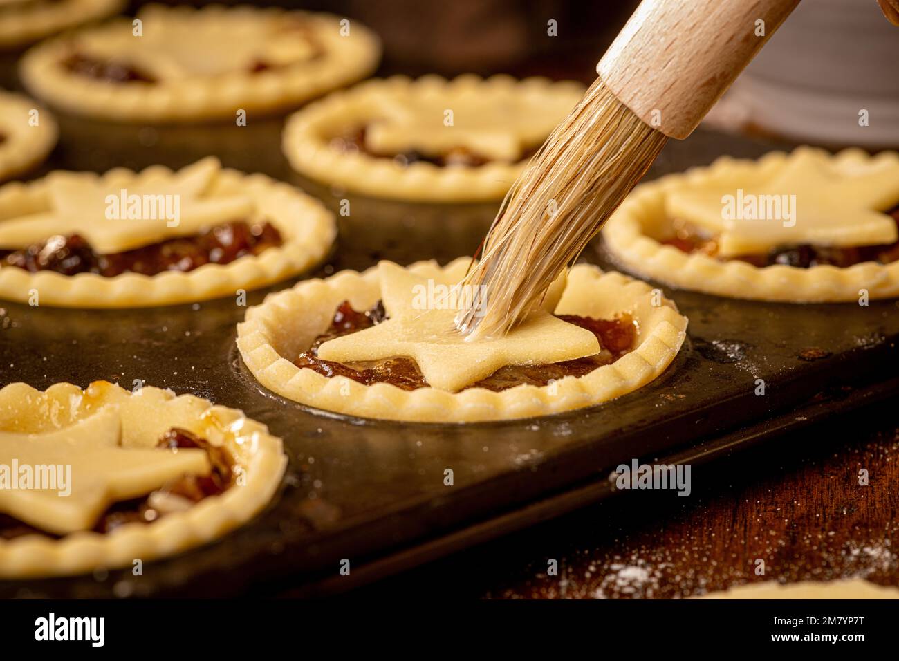 Closeup of star topped mince pies in a dark metal bun tin being brushed