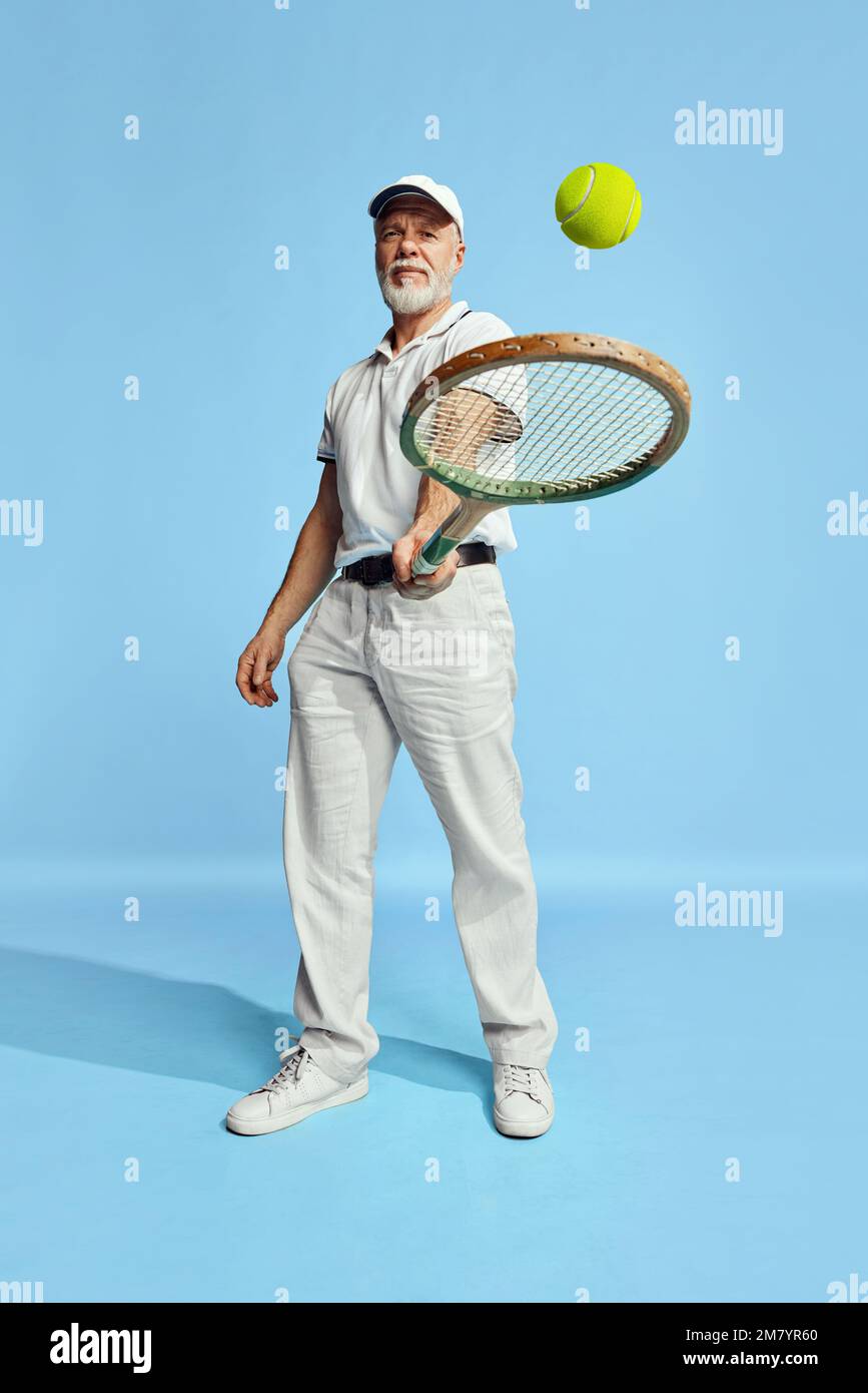 Portrait of handsome senior man in stylish white outfit hitting ball with tennis racket over blue background. Concept of leisure activity, hobby Stock Photo