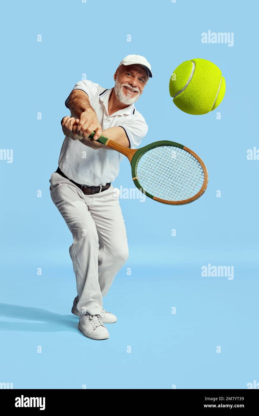 Returning ball. Portrait of handsome senior man in stylish white outfit playing tennis over blue background. Concept of leisure activity, hobby Stock Photo
